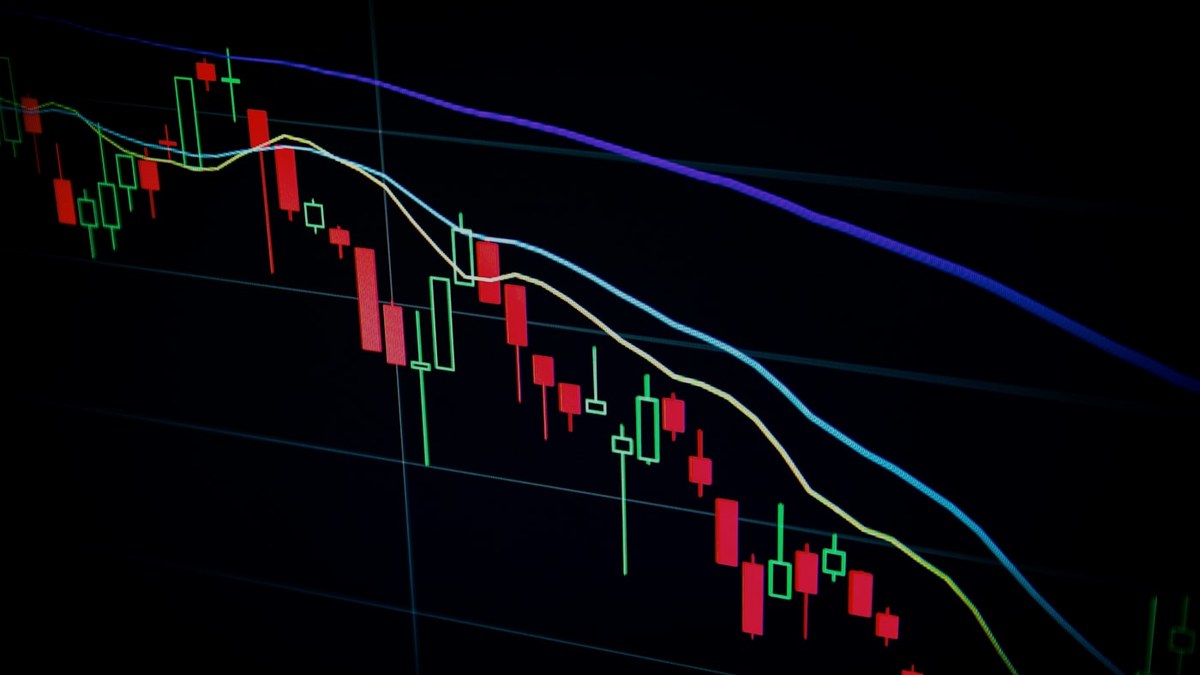 A stock market chart on a dark screen showing red candlestick patterns and falling moving averages.