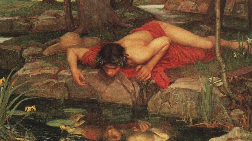 A young man lying on stones, looking at his reflection in a pond.